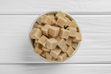 Brown sugar cubes in bowl on white wooden table, top view