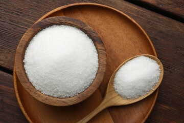 Granulated sugar in bowl and spoon on wooden table, top view