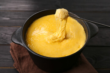 Dipping piece of ham into fondue pot with melted cheese on black wooden table, closeup