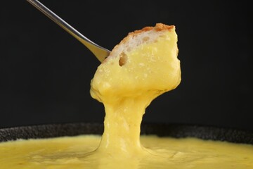 Dipping piece of bread into fondue pot with tasty melted cheese against dark gray background,...