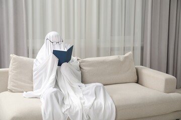 Creepy ghost. Person covered with white sheet reading book on sofa at home, space for text