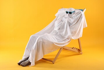 Person in ghost costume and sunglasses relaxing on deckchair against yellow background