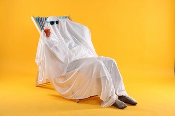 Person in ghost costume and sunglasses with glass of drink relaxing on deckchair against yellow...