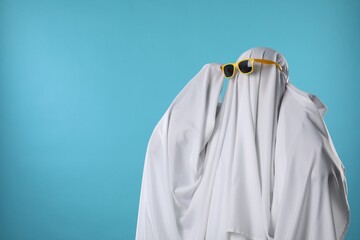 Stylish ghost. Person covered with white sheet in sunglasses on light blue background, space for...