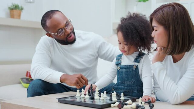 Little kid girl playing chess board at home. Adorable multiracial daughter in living room, enjoy playing piece of chess with parent. Joyful leisure childhood