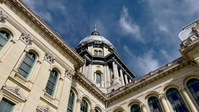 Rear views of the Illinois State Capitol Building in Springfield, Illinois, USA. Cloudy blue skies overhead. The US flag, State of Illinois Flag, and Black History Month Flag fly atop the dome.