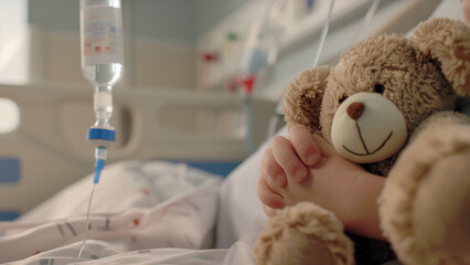 Close-up of young child's hand clutching a bear doll on bed in pediatrics ward with an intravenous drip visible in the background - obrazy, fototapety, plakaty