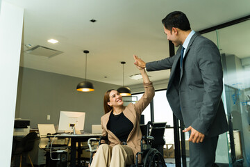 Woman in a wheelchair giving a high five to a colleague in the office - obrazy, fototapety, plakaty