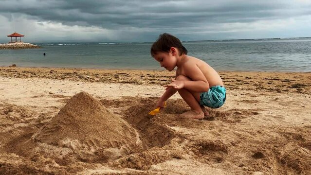 A cute boy is building a sand castle on the shore of the blue sea in cloudy weather. Bad travel weather, clouds and rains. High quality FullHD footage