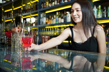 Young professional Asian bartender works behind the bar counter delivering red wine glasses to customers in a nightclub.