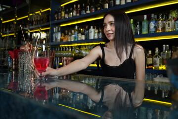 Young professional Asian bartender works behind the bar counter delivering red wine glasses to...