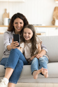 Vertical shot young Hispanic mom and daughter take selfie photos on mobile phone camera, resting on sofa, laughing, hugging, making video call, enjoy new application or game, spend leisure on internet