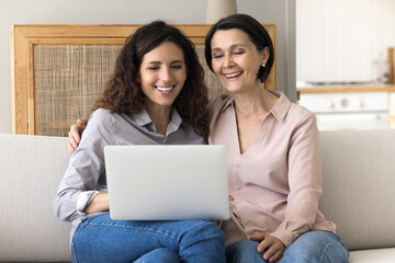Cheerful mother her millennial daughter spend free time together resting on sofa with laptop, watch comedy movie, enjoy videos, buy online. Modern tech usage, subscription services for fun and leisure