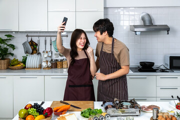 Portrait of young Asian couple male man female woman cheerful standing and preparing food for meal, enjoy cooking, in kitchen at home. Taking selfie.