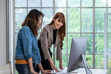 Two smiling young Asian female woman businessperson graphic designer standing and working together, meeting and brainstorming, using computer in office.