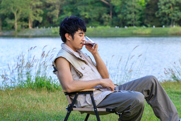 Young Asian male man sitting on chair, drinking coffee, feeling relax and raising hands, during camping beside lake and mountain.