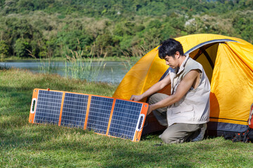 Young Asian male man sitting in front of camping tent beside lake and mountain with solar cell, charging mobile phone.