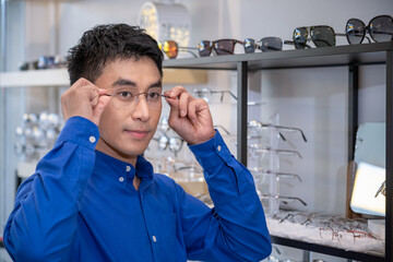 Portrait of Young Asian male man choosing glasses in optical shop, trying eyeglasses. Eyecare concept.