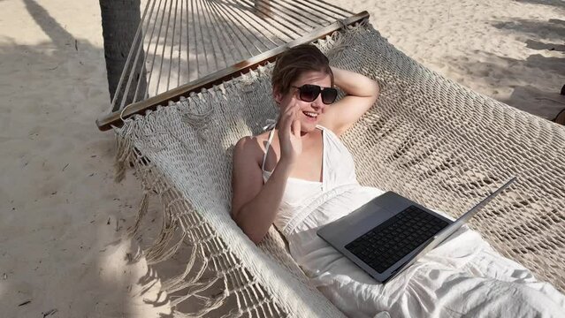Relaxed adult person working on a laptop while lounging on a hammock at a sandy beach, depicting remote work or digital nomad lifestyle