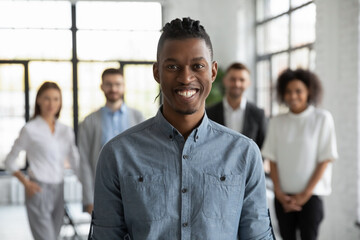 Young African company employee in foreground, with his colleagues behind, smiling and looking at camera in modern office. Happy business trainer or start-up founder leading team of workers to success - obrazy, fototapety, plakaty