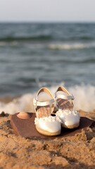 First communion slippers on a stone in the sand of a beach with the blue sea in the background.