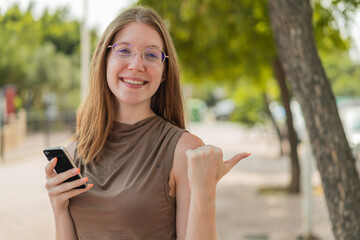 French girl with glasses using mobile phone at outdoors pointing to the side to present a product