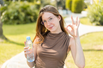 French girl with glasses holding a bottle of water at outdoors showing ok sign with fingers