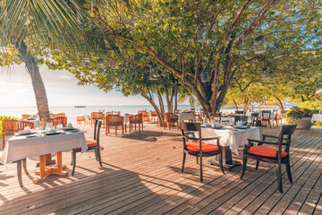 Outdoor restaurant beach bar. Luxury table setting tropical beach restaurant. Sunset light trees wooden tables chairs under beautiful romantic vintage sky, sea view. Cozy hotel resort tourism travel