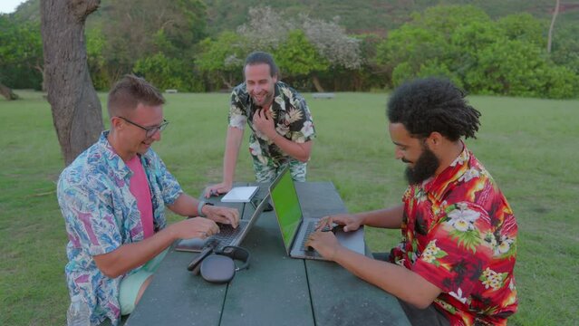 Three men enjoying leisure and work, sitting at a table with green screen laptop at park chatting and laughing with laptops. African American male and caucasian mens wearing Hawaiian shirts 