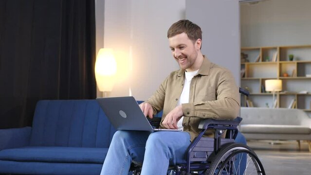 Handsome man with a disability sitting in wheelchair with laptop on knees and looking at camera. Caucasian male in casual wear having opportunities to remote work at home