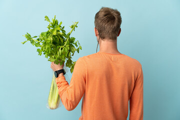 Young blonde man holding a celery isolated on blue background in back position