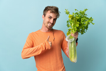 Young blonde man holding a celery isolated on blue background with thumbs up because something good...