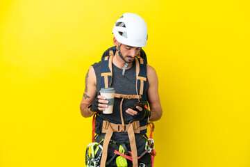 Young caucasian rock climber man isolated on yellow background holding coffee to take away and a mobile