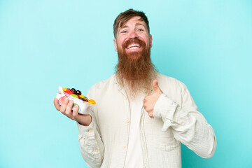 Redhead man with long beard holding a bowl of fruit isolated on blue background with thumbs up...