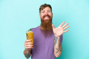 Redhead man with long beard holding a cocktail isolated on blue background saluting with hand with...