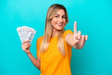 Young Uruguayan woman taking a lot of money isolated on blue background showing and lifting a finger