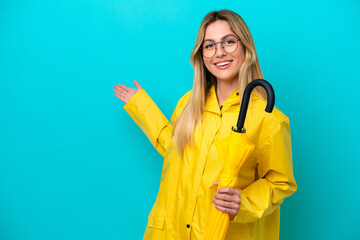 Young Uruguayan woman with rainproof coat and umbrella isolated on blue background extending hands...