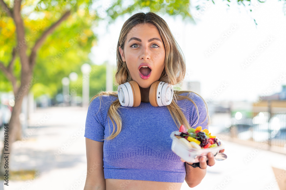 Wall mural Young pretty sport woman holding a bowl of fruit at outdoors with surprise and shocked facial expression