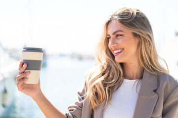 Pretty blonde Uruguayan woman holding a take away coffee at outdoors with happy expression