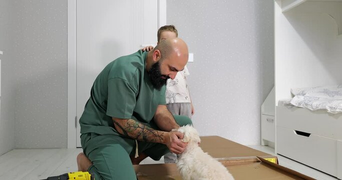Concept of happy fatherhood. A bearded male owner play with his cute fluffy white puppy at home, while the wardrobe is being assembled in the child room with a son on the background.