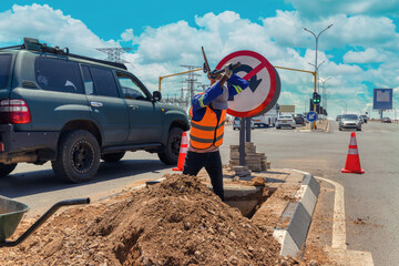 african american worker with a pickaxe digging a ditch in the highway to repair cabling on an isle at an intersection - obrazy, fototapety, plakaty