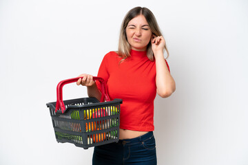 Young Rumanian woman holding a shopping basket full of food isolated on white background frustrated and covering ears