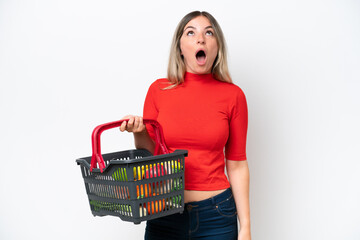 Young Rumanian woman holding a shopping basket full of food isolated on white background looking up and with surprised expression