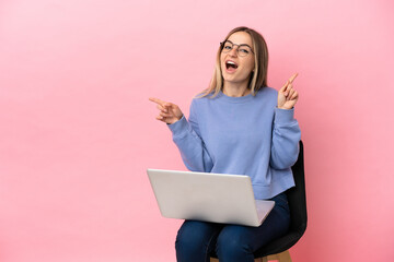 Young woman sitting on a chair with laptop over isolated pink background pointing finger to the...
