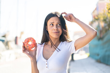 Young woman at outdoors holding a donut at outdoors having doubts and with confuse face expression