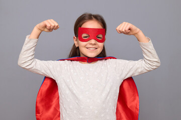 Strong powerful little girl wearing superhero costume and mask isolated over gray background standing with raised arms showing her biceps looking smiling at camera - Powered by Adobe
