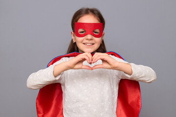 Delighted charming cute little girl wearing superhero costume and mask isolated over gray background looking smiling at camera showing heart shape with hands love symbol