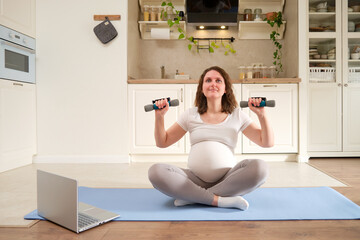 Pregnant woman is engaged in strength training with dumbbells in the home kitchen, exercises to...