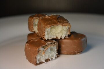 Pieces, slices of sweet coconut bar covered with milk chocolate placed on a white glass plate.