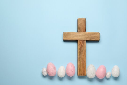 Wooden cross and painted Easter eggs on light blue background, flat lay. Space for text